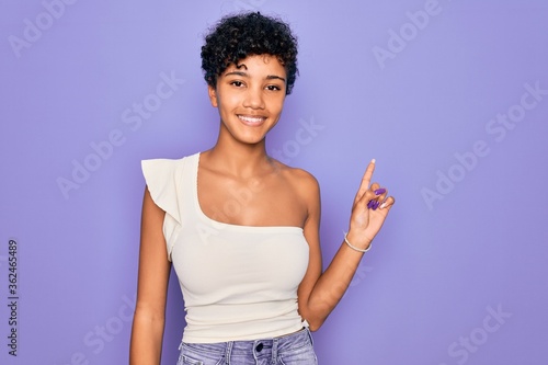 Young beautiful african american afro woman wearing casual t-shirt over purple background with a big smile on face, pointing with hand finger to the side looking at the camera.