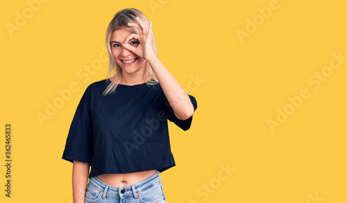 Young beautiful blonde woman wearing casual t-shirt doing ok gesture with hand smiling, eye looking through fingers with happy face.