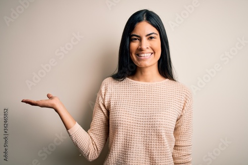 Young beautiful hispanic woman wearing elegant pink sweater over isolated background smiling cheerful presenting and pointing with palm of hand looking at the camera.