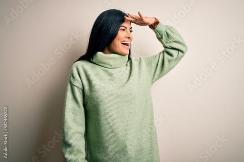 Young beautiful hispanic woman wearing green winter sweater over isolated background very happy and smiling looking far away with hand over head. Searching concept. © Krakenimages.com
