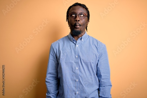 Young handsome african american man wearing shirt and glasses over yellow background making fish face with lips, crazy and comical gesture. Funny expression.