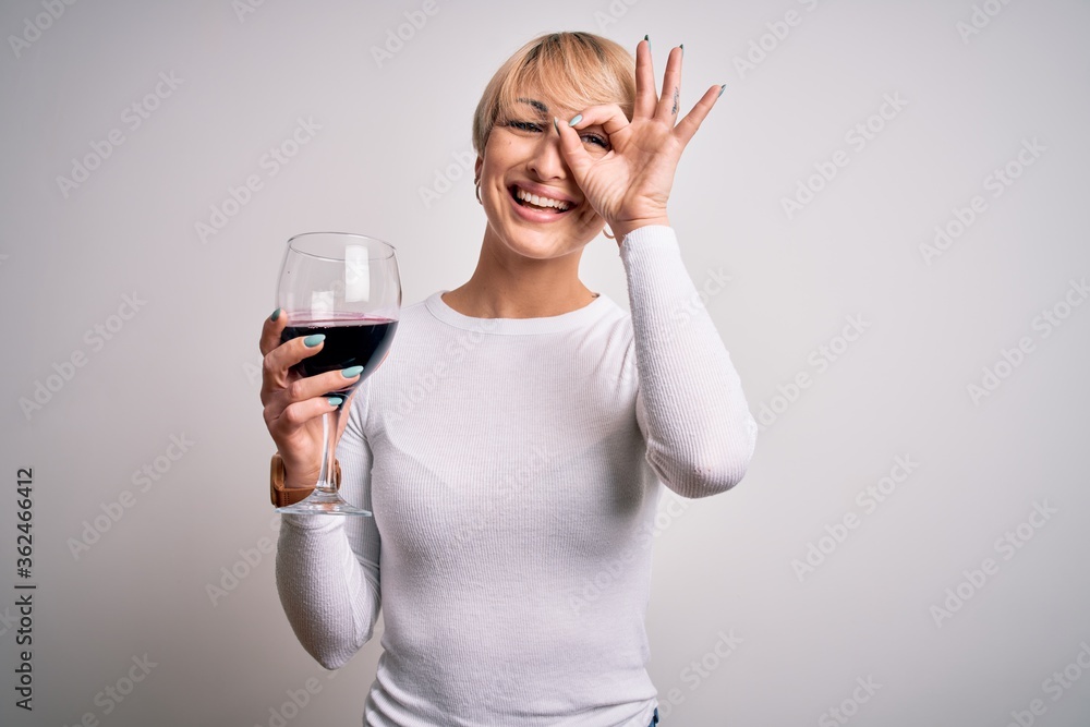 Young blonde woman with short hair drinking a glass of red wine over isolated background with happy face smiling doing ok sign with hand on eye looking through fingers Young blonde woman with short hair drinking a glass of red wine over isolated background with happy face smiling doing ok sign with hand on eye looking through fingers