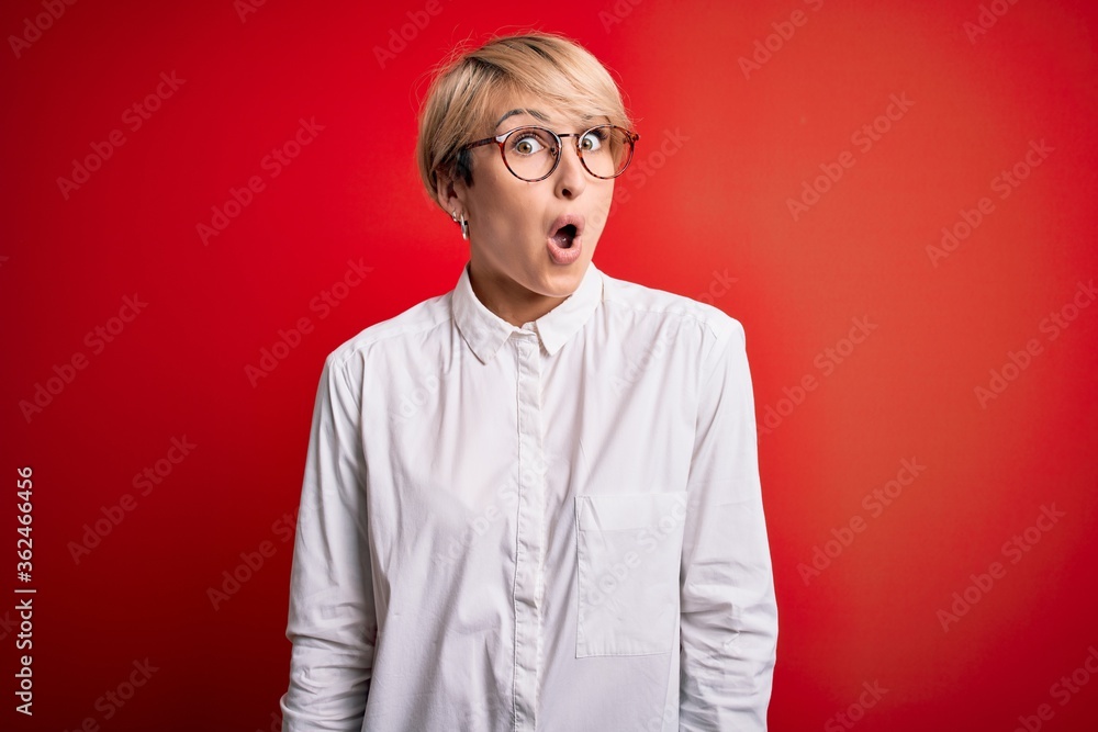 Young blonde business woman with short hair wearing glasses over red background afraid and shocked with surprise expression, fear and excited face. Young blonde business woman with short hair wearing glasses over red background afraid and shocked with surprise expression, fear and excited face.