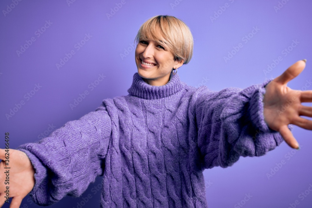 Young blonde woman with short hair wearing winter turtleneck sweater over purple background looking at the camera smiling with open arms for hug. Cheerful expression embracing happiness. Young blonde woman with short hair wearing winter turtleneck sweater over purple background looking at the camera smiling with open arms for hug. Cheerful expression embracing happiness.