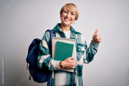 Young blonde student woman with short hair wearing backpack and holding university books happy with big smile doing ok sign, thumb up with fingers, excellent sign