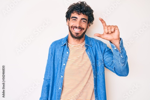 Handsome young man with curly hair and bear wearing casual denim jacket smiling and confident gesturing with hand doing small size sign with fingers looking and the camera. measure concept.