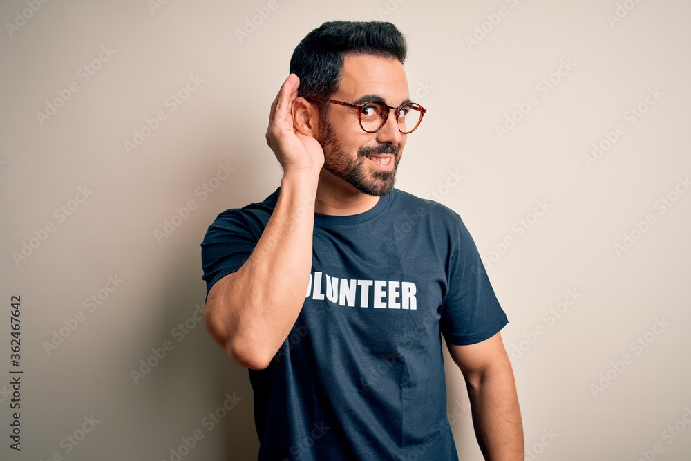 Handsome man with beard wearing t-shirt with volunteer message over white background smiling with hand over ear listening an hearing to rumor or gossip. Deafness concept. Handsome man with beard wearing t-shirt with volunteer message over white background smiling with hand over ear listening an hearing to rumor or gossip. Deafness concept.