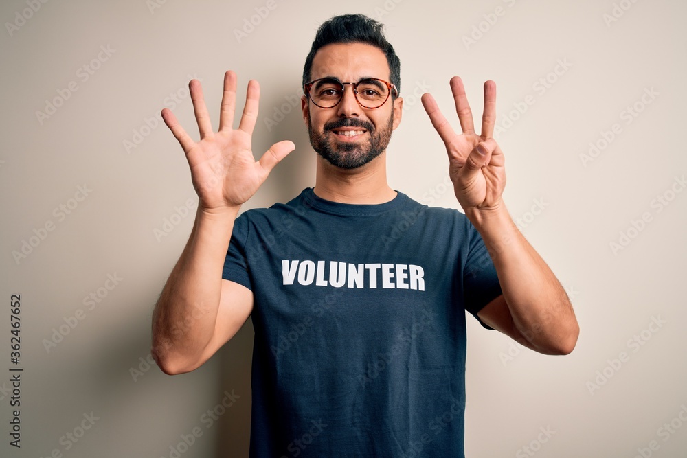 Handsome man with beard wearing t-shirt with volunteer message over white background showing and pointing up with fingers number eight while smiling confident and happy. Handsome man with beard wearing t-shirt with volunteer message over white background showing and pointing up with fingers number eight while smiling confident and happy.