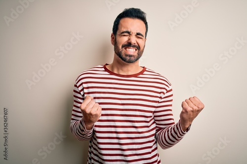 Young handsome man with beard wearing casual striped t-shirt standing over white background excited for success with arms raised and eyes closed celebrating victory smiling. Winner concept.