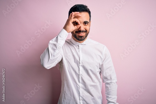 Young handsome man with beard wearing casual shirt standing over pink background doing ok gesture with hand smiling, eye looking through fingers with happy face.
