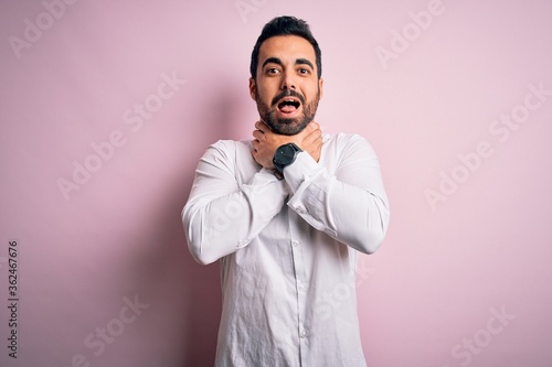 Young handsome man with beard wearing casual shirt standing over pink background shouting and suffocate because painful strangle. Health problem. Asphyxiate and suicide concept.