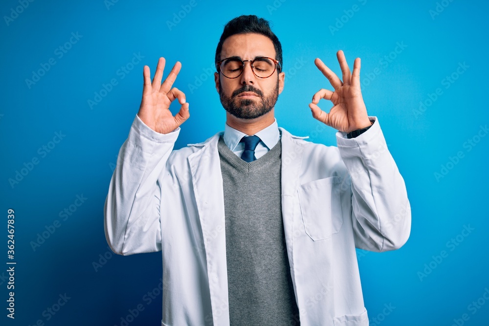 Young handsome doctor man with beard wearing coat and glasses over blue background relax and smiling with eyes closed doing meditation gesture with fingers. Yoga concept. Young handsome doctor man with beard wearing coat and glasses over blue background relax and smiling with eyes closed doing meditation gesture with fingers. Yoga concept.