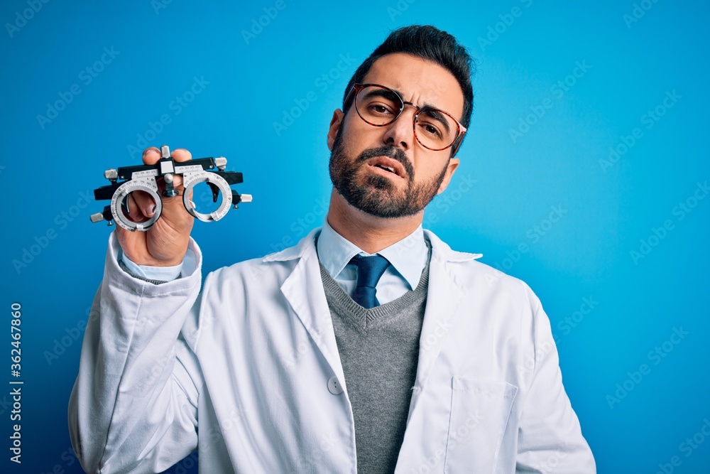 Young handsome optical man with beard holding optometry glasses over blue background looking sleepy and tired, exhausted for fatigue and hangover, lazy eyes in the morning.