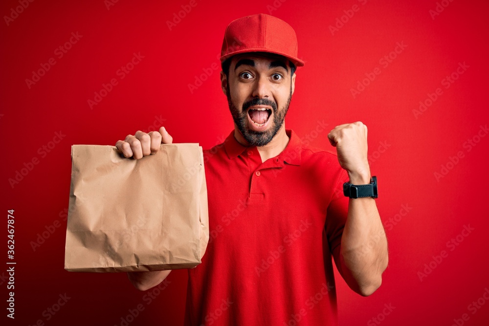 Young handsome delivery man with beard wearing cap holding takeaway paper bag with food screaming proud and celebrating victory and success very excited, cheering emotion