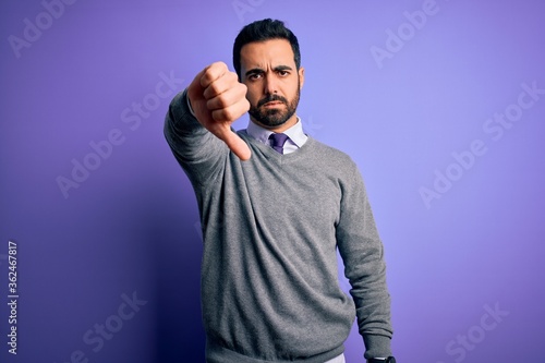 Handsome businessman with beard wearing casual tie standing over purple background looking unhappy and angry showing rejection and negative with thumbs down gesture. Bad expression.