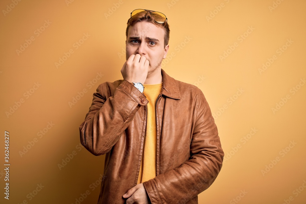 Young handsome redhead man wearing casual leather jacket over isolated yellow background looking stressed and nervous with hands on mouth biting nails. Anxiety problem. Young handsome redhead man wearing casual leather jacket over isolated yellow background looking stressed and nervous with hands on mouth biting nails. Anxiety problem.