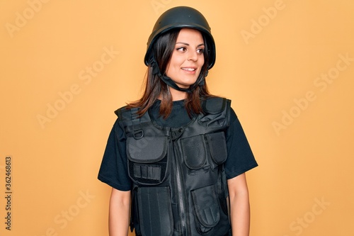 Принти на полотні Young beautiful brunette soldier woman wearing bulletproof and security helmet looking away to side with smile on face, natural expression