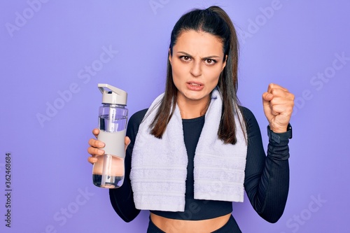 Young beautiful brunette sporty woman using sport towel drinking bottle of water annoyed and frustrated shouting with anger, yelling crazy with anger and hand raised © Krakenimages.com
