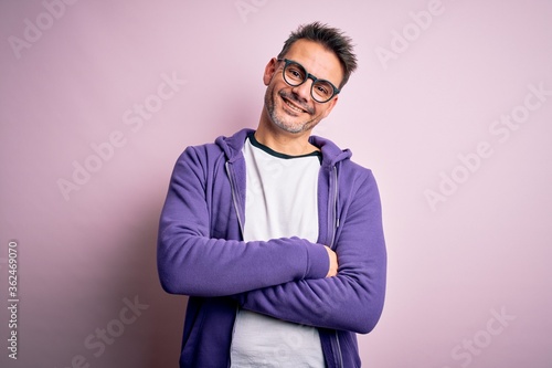 Young handsome man wearing purple sweatshirt and glasses standing over pink background happy face smiling with crossed arms looking at the camera. Positive person.