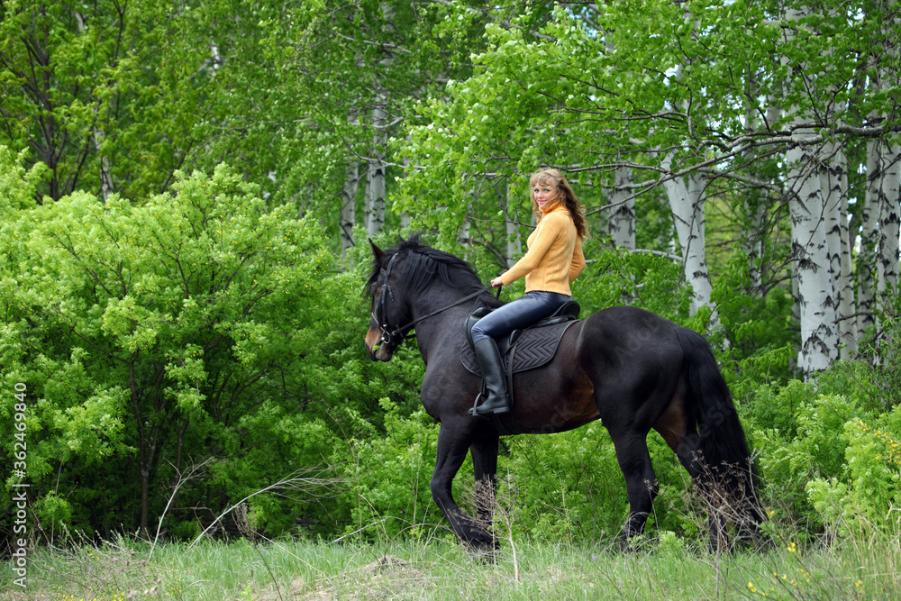 Beautiful blond girl with black horse in summer forest. Mare, animal