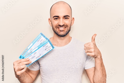 Young handsome bald man holding airline boarding pass smiling happy and positive, thumb up doing excellent and approval sign photo