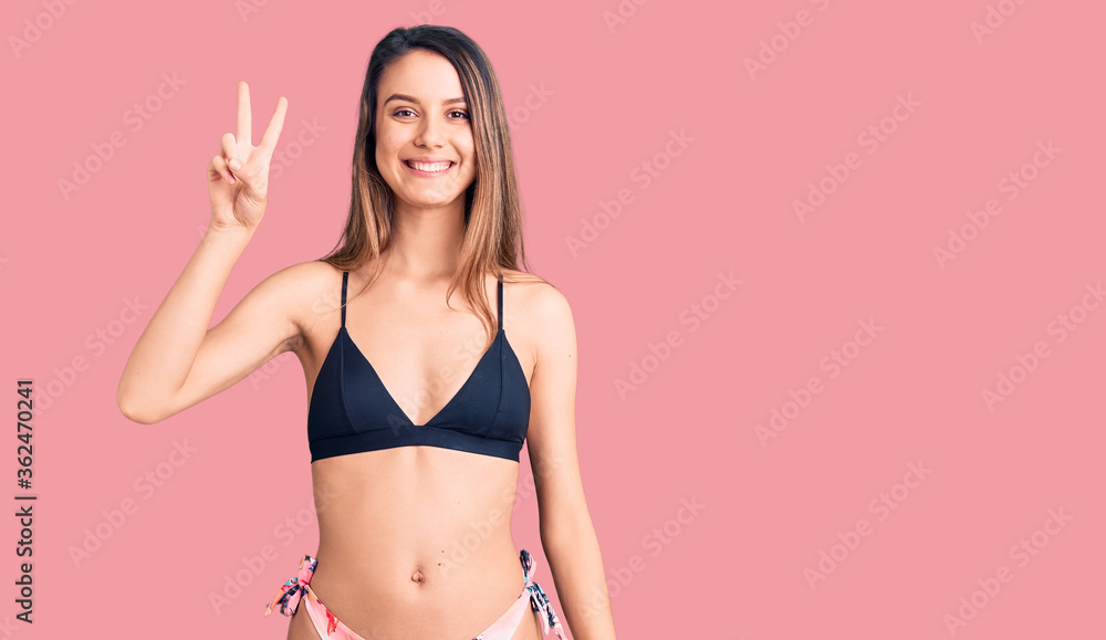 Naklejka premium Young beautiful girl wearing bikini smiling with happy face winking at the camera doing victory sign. number two.
