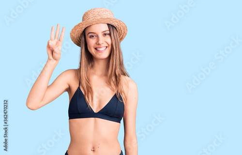 Young beautiful girl wearing bikini and hat showing and pointing up with fingers number three while smiling confident and happy.
