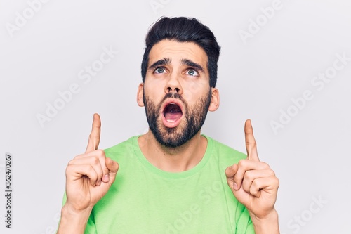 Young handsome man with beard wearing casual t-shirt amazed and surprised looking up and pointing with fingers and raised arms.
