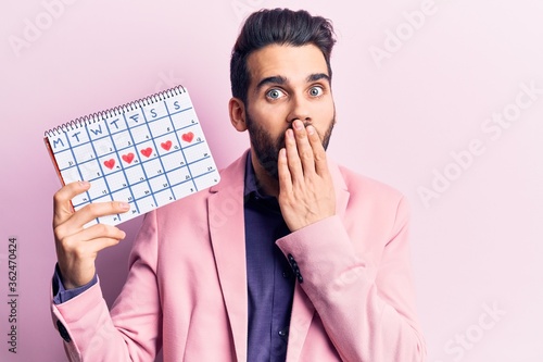 Young handsome man with beard holding calendar with hearts covering mouth with hand, shocked and afraid for mistake. surprised expression photo