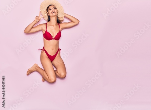 Young beautiful girl on vacation wearing bikini and summer hat smiling happy. Jumping with smile on face doing victory sign over isolated pink background