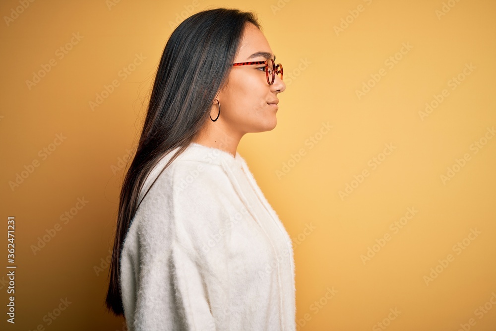 Young beautiful asian woman wearing casual sweater and glasses over yellow background looking to side, relax profile pose with natural face with confident smile. Young beautiful asian woman wearing casual sweater and glasses over yellow background looking to side, relax profile pose with natural face with confident smile.