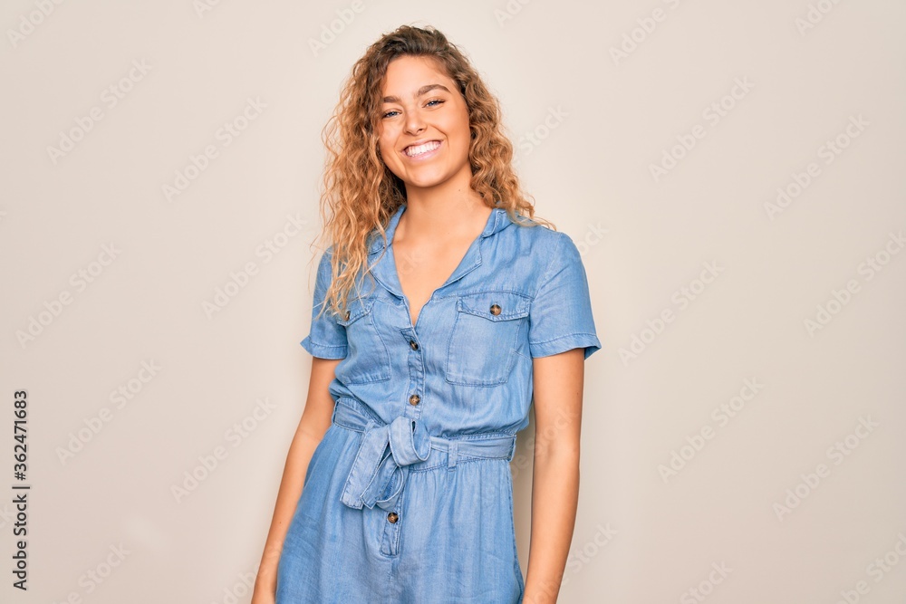 Young beautiful woman with blue eyes wearing casual denim dress over white background looking away to side with smile on face, natural expression. Laughing confident. Young beautiful woman with blue eyes wearing casual denim dress over white background looking away to side with smile on face, natural expression. Laughing confident.