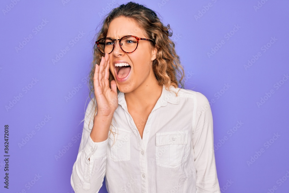 Young beautiful woman with blue eyes wearing casual shirt and glasses over purple background shouting and screaming loud to side with hand on mouth. Communication concept.