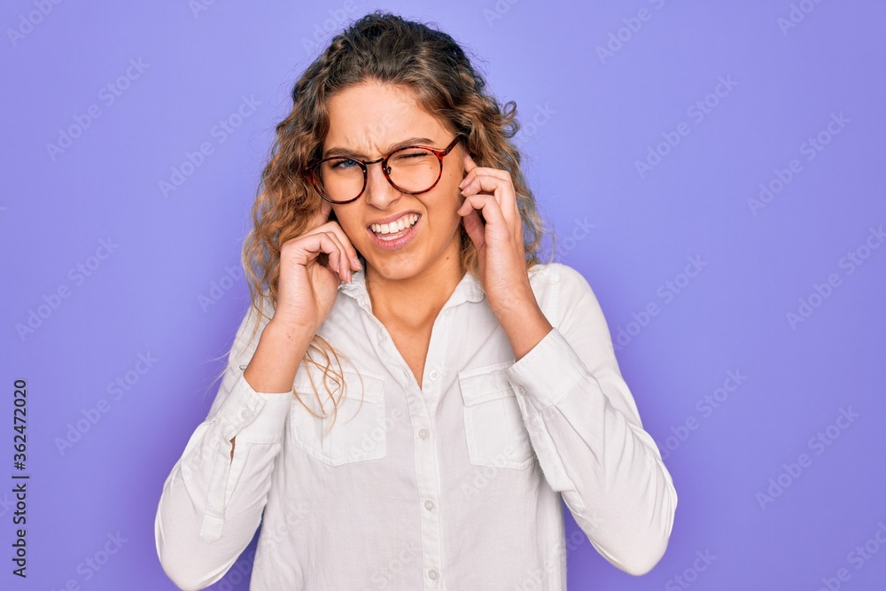 Young beautiful woman with blue eyes wearing casual shirt and glasses over purple background covering ears with fingers with annoyed expression for the noise of loud music. Deaf concept. Young beautiful woman with blue eyes wearing casual shirt and glasses over purple background covering ears with fingers with annoyed expression for the noise of loud music. Deaf concept.