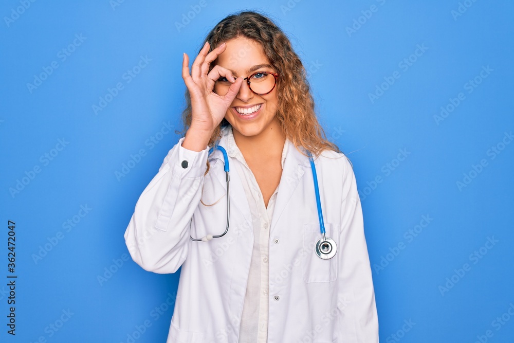 Young beautiful blonde doctor woman with blue eyes wearing coat and stethoscope doing ok gesture with hand smiling, eye looking through fingers with happy face. Young beautiful blonde doctor woman with blue eyes wearing coat and stethoscope doing ok gesture with hand smiling, eye looking through fingers with happy face.