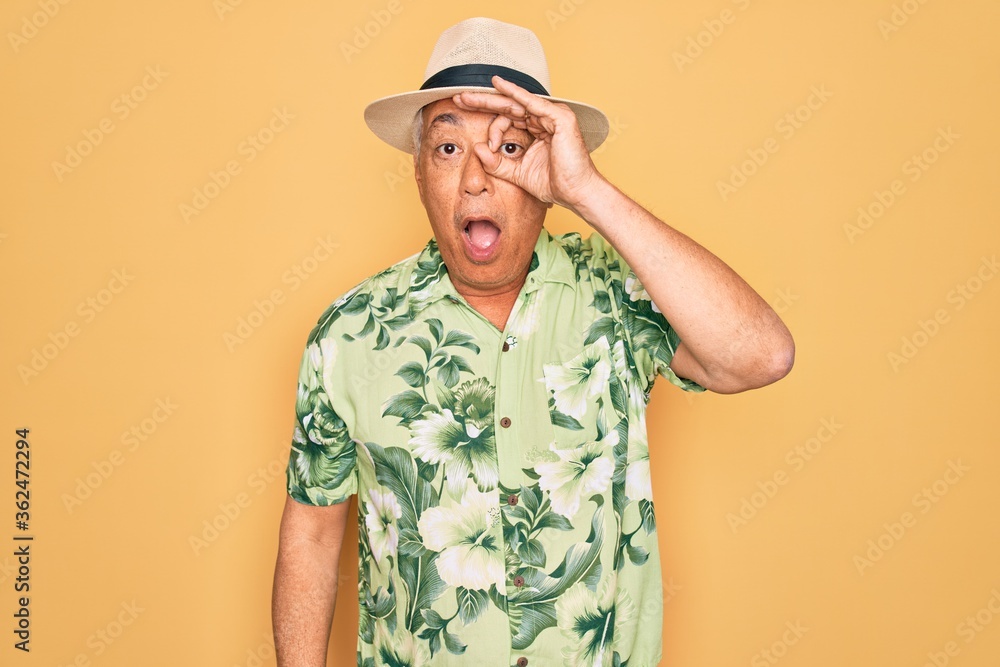 Middle age senior grey-haired man wearing summer hat and floral shirt on beach vacation doing ok gesture shocked with surprised face, eye looking through fingers. Unbelieving expression.