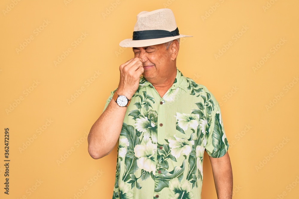 Middle age senior grey-haired man wearing summer hat and floral shirt on beach vacation smelling something stinky and disgusting, intolerable smell, holding breath with fingers on nose. Bad smell Middle age senior grey-haired man wearing summer hat and floral shirt on beach vacation smelling something stinky and disgusting, intolerable smell, holding breath with fingers on nose. Bad smell