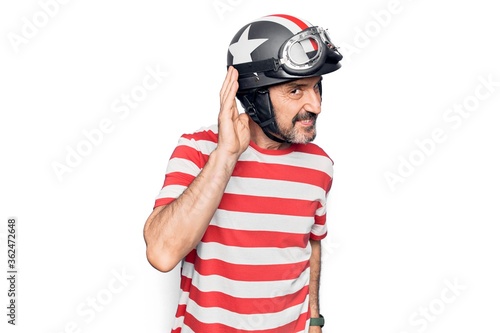Middle age handsome motorcyclist man wearing moto helmet over isolated white background smiling with hand over ear listening and hearing to rumor or gossip. Deafness concept.