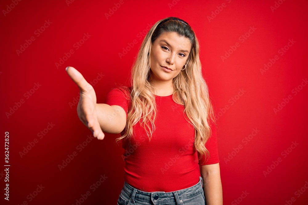 Young beautiful blonde woman wearing casual t-shirt standing over isolated red background smiling cheerful offering palm hand giving assistance and acceptance. Young beautiful blonde woman wearing casual t-shirt standing over isolated red background smiling cheerful offering palm hand giving assistance and acceptance.