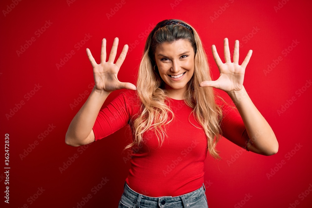 Young beautiful blonde woman wearing casual t-shirt standing over isolated red background showing and pointing up with fingers number ten while smiling confident and happy. Young beautiful blonde woman wearing casual t-shirt standing over isolated red background showing and pointing up with fingers number ten while smiling confident and happy.