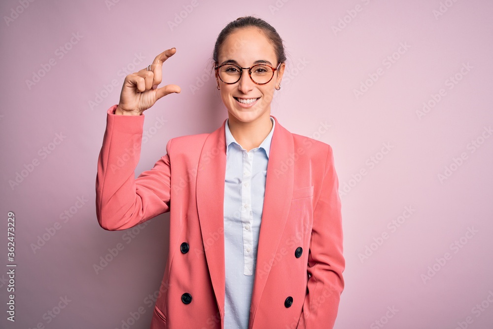 Young beautiful businesswoman wearing jacket and glasses over isolated pink background smiling and confident gesturing with hand doing small size sign with fingers looking and the camera. Measure. Young beautiful businesswoman wearing jacket and glasses over isolated pink background smiling and confident gesturing with hand doing small size sign with fingers looking and the camera. Measure.