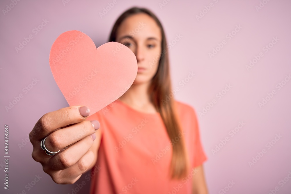 Young beautiful romantic woman holding paper heart shape over isolated pink background with a confident expression on smart face thinking serious Young beautiful romantic woman holding paper heart shape over isolated pink background with a confident expression on smart face thinking serious