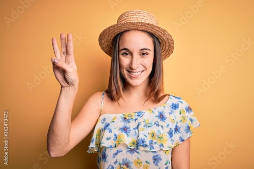 Young beautiful woman wearing casual t-shirt and summer hat over isolated yellow background showing and pointing up with fingers number three while smiling confident and happy.