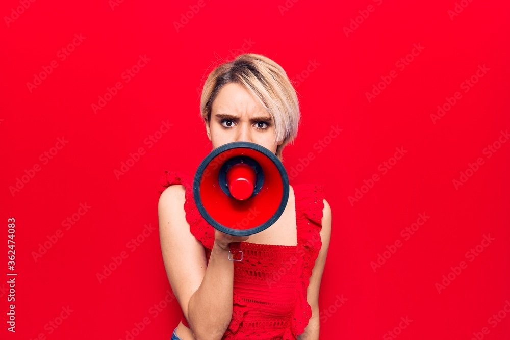 Fototapeta premium Young beautiful blonde woman with angry expression. Screaming loud using megaphone standing over isolated red background