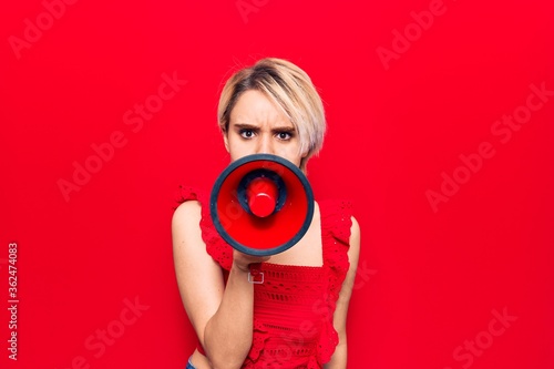 Young beautiful blonde woman with angry expression. Screaming loud using megaphone standing over isolated red background