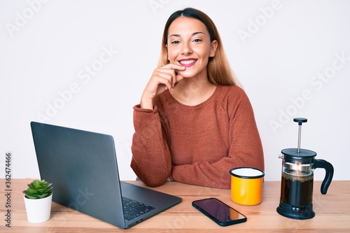 Young brunette woman working at the office drinking a cup of coffee smiling looking confident at the camera with crossed arms and hand on chin. thinking positive.