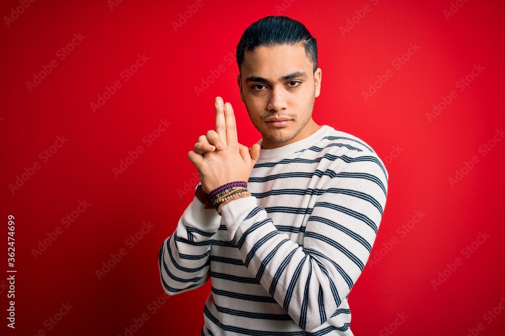 Young brazilian man wearing casual striped t-shirt standing over isolated red background Holding symbolic gun with hand gesture, playing killing shooting weapons, angry face Young brazilian man wearing casual striped t-shirt standing over isolated red background Holding symbolic gun with hand gesture, playing killing shooting weapons, angry face