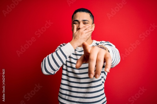 Young brazilian man wearing casual striped t-shirt standing over isolated red background laughing at you, pointing finger to the camera with hand over mouth, shame expression