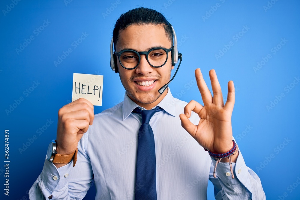 Young brazilian call center agent man overworked holding reminder paper with help message doing ok sign with fingers, excellent symbol