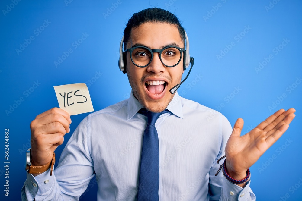 Young brazilian call center agent man holding reminder paper with yes message very happy and excited, winner expression celebrating victory screaming with big smile and raised hands Young brazilian call center agent man holding reminder paper with yes message very happy and excited, winner expression celebrating victory screaming with big smile and raised hands
